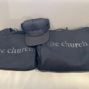 May include: Black apparel set featuring the text "the church." Includes a black crew neck t-shirt, a black hoodie, and a black trucker hat. The hat has a mesh back and the text is in a tone-on-tone design.