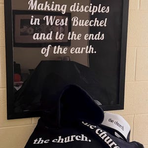 May include: A black hoodie, a black cap, and a white and black cap are displayed. The hoodie and cap feature the text "the church." in white. Behind the items is a framed print with the words "the church. Making disciples in West Buechel and to the ends of the earth."
