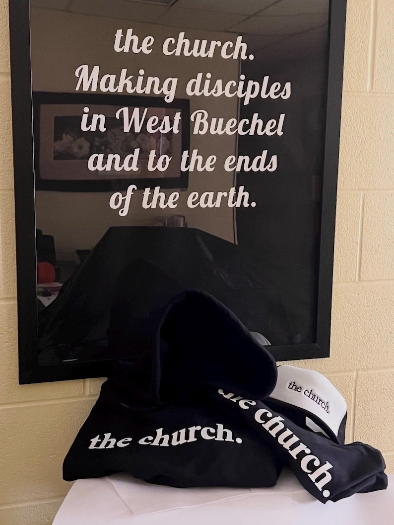 May include: A black hoodie, a black cap, and a white and black cap are displayed. The hoodie and cap feature the text "the church." in white. Behind the items is a framed print with the words "the church. Making disciples in West Buechel and to the ends of the earth."