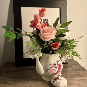 May include: A white teapot with a floral design, filled with pink, red, and white flowers and greenery. The lid is in front. A framed print is in the background. The teapot is a decorative item.