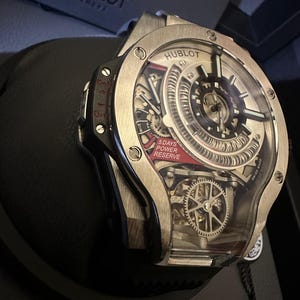 Op de afbeelding: Een zilveren Hublot-horloge met een transparante wijzerplaat, met ingewikkelde interne mechanismen. Het horloge heeft een rode accentkleur met de tekst "5 DAYS POWER RESERVE". Het horloge ligt op een zwarte ondergrond.