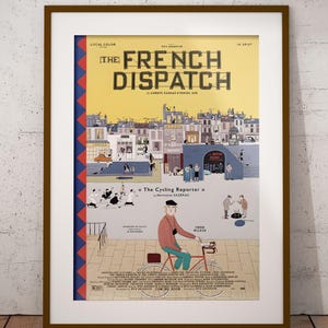 May include: Framed poster for "The French Dispatch" film. The poster features a stylized illustration of a city scene with the film's title in large letters. A man on a bicycle is in the foreground. The poster is in a brown frame.