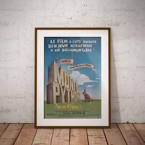 May include: Framed vintage movie poster for "Monty Python and the Holy Grail." The poster features a castle, a rabbit, and text in French. The poster is in a brown frame and is displayed against a white wall and wooden floor.
