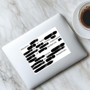 May include: White sticker on a laptop with the text "Subject: go back to sleep, compliance is mandatory, truth is dangerous." The text is partially obscured by black bars. A cup of coffee sits to the right.