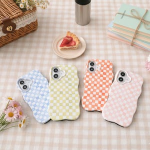 May include: Four colorful phone cases with a wavy edge, each featuring a checkered pattern in blue, yellow, orange, and pink. The cases are arranged on a checkered tablecloth, alongside a pie, a basket, and a stack of books.