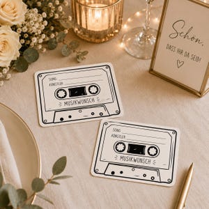 Op de afbeelding: Twee zwart-witte onderzetters in de vorm van een cassettebandje met de tekst "SONG", "KÜNSTLER:" en "MUSIKWUNSCH" erop gedrukt. Een goudkleurige pen, een kaars in een glazen houder en een bord met de tekst "Schön, dass ihr da seid!" zijn ook zichtbaar.