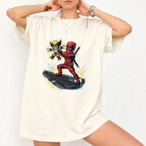 Puede incluir: Camiseta color crema con un gr&aacute;fico colorido de Deadpool y Wolverine. Deadpool, con su traje rojo y negro, est&aacute; de pie sobre una plataforma oscura, sosteniendo a Wolverine. Wolverine est&aacute; con su traje amarillo y azul. El dise&ntilde;o es de estilo caricatura.