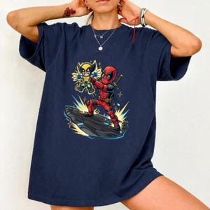 Puede incluir: Camiseta azul marino con una ilustraci&oacute;n de dibujos animados de Deadpool y Wolverine. Deadpool lleva un traje rojo y negro, y Wolverine un disfraz amarillo y azul. El gr&aacute;fico est&aacute; sobre una roca negra con efectos de estrellas.