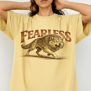 May include: A light yellow t-shirt featuring the word "FEARLESS" in brown, arched lettering above a detailed illustration of a snarling wolf. The wolf is brown and tan, with its mouth open, and is in a running pose.