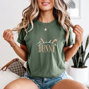 Può includere: T-shirt verde oliva con la scritta "Teacher Bunny" in corsivo e grafica di orecchie di coniglio. La maglietta è a maniche corte, in stile casual.