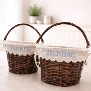 以下が含まれることがあります： アーチ型のハンドルが付いた、2つの茶色の籐かご。各バスケットには、スカラップエッジのクリーム色の生地の裏地が付いています。1つのバスケットには青い文字で「PARKER」と書かれており、もう1つには「BECKHAM」と書かれています。