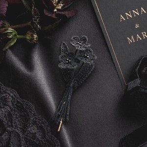 Könnte beinhalten: Ein schwarzes gehäkeltes Blumenbouquet mit einer goldenen Nadel, das auf einer dunklen Oberfläche liegt. Das Bouquet hat drei Blumen mit Perlen in der Mitte. Eine schwarze Einladungskarte mit goldenem Text ist im Hintergrund.
