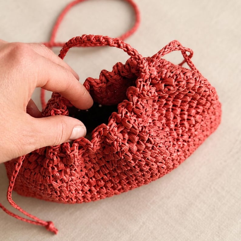 May include: A small, woven red bag with a drawstring closure and a handle. The bag is made of a textured material and has a rounded shape. The bag is held open, revealing a dark interior.