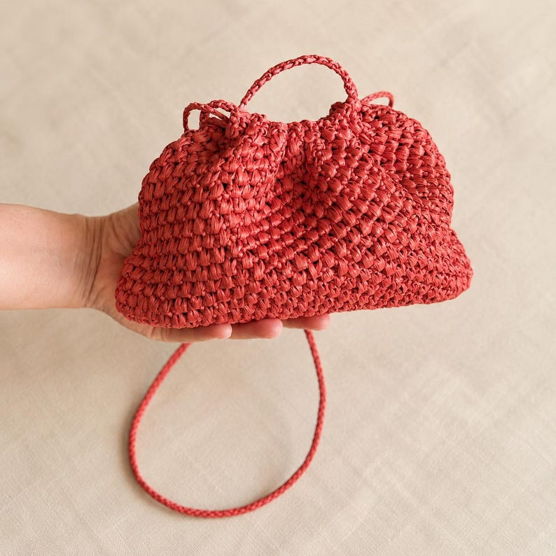 May include: A red woven handbag with a top handle and a long, thin strap. The bag is held in a hand, showcasing its texture and shape. The bag's color is a vibrant red, and the weave pattern is visible.