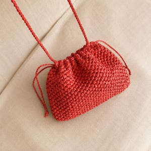 May include: A red woven drawstring bag with a shoulder strap. The bag is made of a textured material and has a rounded shape. The drawstring closure is visible at the top of the bag.