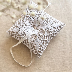 May include: A square, off-white crochet ring pillow with two silver rings. The pillow has a beige fabric base and is adorned with a white ribbon. The crochet design is intricate and detailed, perfect for a wedding.