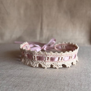 Può includere: Un girocollo all'uncinetto color crema con un nastro di raso rosa infilato nel design. Il girocollo presenta un bordo smerlato e un fiocco di raso rosa sul retro. L'accessorio è progettato per essere indossato intorno al collo.