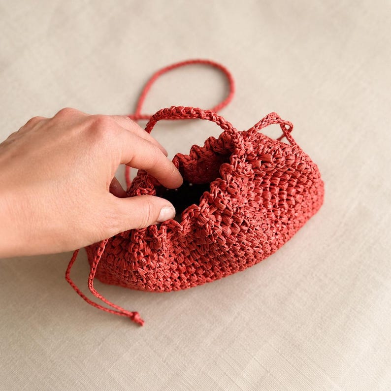 May include: A small, red woven drawstring bag with a matching handle and drawstring closure. The bag is made of a textured material and has a gathered top. The bag is held open by a hand.