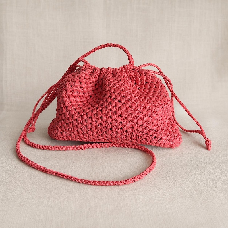 May include: A coral-colored woven raffia handbag with a drawstring closure and a braided shoulder strap. The bag has a rounded shape and a textured surface. The strap is long enough to be worn over the shoulder.