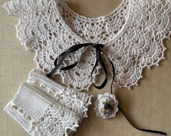 May include: A white crochet collar, cuffs, and a small flower pin with a black ribbon. The collar and cuffs have a scalloped edge and intricate openwork design. The cuffs are adorned with small, white beads.