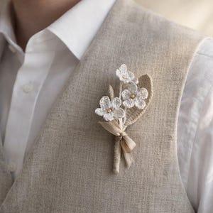 May include: A white crochet flower boutonniere with three flowers and a leaf, accented with small gold beads. The boutonniere is tied with a beige ribbon and is pinned to a light tan waistcoat over a white shirt.