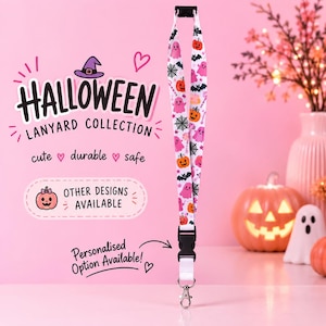 May include: A white lanyard featuring a repeating Halloween pattern of orange pumpkins, pink ghosts, black bats, and spiderwebs. The strap includes a black plastic safety breakaway buckle at the top and a black side-release buckle with a metal lobster clasp.