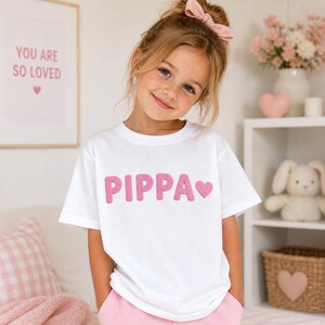 May include: A young girl wearing a white t-shirt with the name "PIPA" in pink letters and a pink heart. The girl is smiling and wearing a pink bow in her hair. The background includes a framed print that says "YOU ARE SO LOVED".