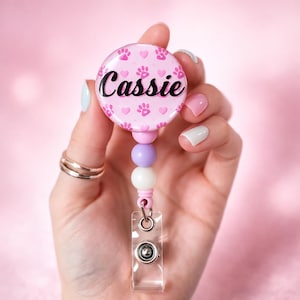 May include: A circular badge reel featuring a light pink background with a repeating pattern of dark pink paw prints and small hearts. The name Cassie is printed in black script across the center. The reel includes a pink, purple, and white bead accent.