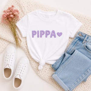 May include: A white t-shirt with the name "PIPPA" in purple, textured letters and a purple heart. The shirt is paired with light blue jeans, white sneakers, and a bouquet of pink flowers, all arranged on a cream-colored knit blanket.