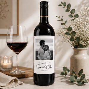 May include: A dark glass wine bottle with a custom white rectangular label featuring a black and white photograph of a couple embracing. The label includes the text Happy Anniversary, the names Sam and Colbie, and the date 01/07/2026 in black font.