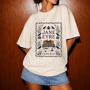 Puede incluir: Una camiseta de manga corta color hueso con un estampado gráfico de la portada del libro Jane Eyre de Charlotte Brontë. El diseño incluye una mansión en llamas, cuervos y bordes florales, con el texto JANE EYRE y Charlotte Brontë.