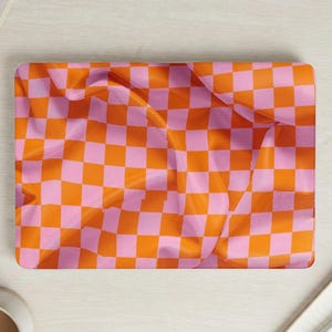 May include: A rectangular, pink and orange checkered pattern. The design is a repeating grid of squares in alternating colors. The fabric appears to be draped, creating soft folds and shadows. The overall aesthetic is bright and playful.