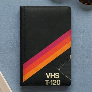 May include: A rectangular black notebook featuring a diagonal stripe pattern in shades of pink, red, and orange. The bottom right corner displays the white text VHS T-120, with a distressed, worn graphic effect near the lower right edge.