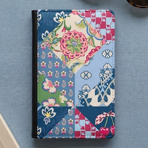 May include: A rectangular notebook with a black spine and a patchwork cover design. The cover features various geometric sections displaying floral patterns, paisley motifs, a red and white checkered pattern with cherries, and blue tones.