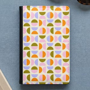 Könnte beinhalten: Ein rechteckiges Notizbuch mit schwarzem Buchrücken und cremefarbenem Einband, das ein sich wiederholendes geometrisches Muster aus Halbkreisen in Orange, Olivgrün, Hellrosa und Kornblumenblau in einem Raster zeigt.