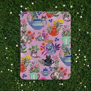 Pode incluir: Um objeto retangular com um design caprichoso de gatos, bules e flores em um fundo rosa. Os gatos são retratados em várias poses, alguns com xícaras de chá, e os bules têm padrões intrincados. As flores são em tons de amarelo, laranja e rosa.
