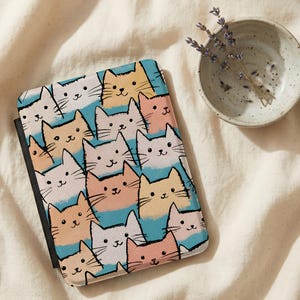 May include: A colorful cat-themed case featuring a repeating pattern of cartoon cat faces in various colors, including peach, yellow, and white, set against a light blue background. The case has a black spine and is likely for an e-reader or tablet.