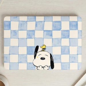 May include: A rectangular laptop skin featuring a blue and white checkered pattern. The design includes a cartoon illustration of Snoopy with Woodstock perched on his head. The overall color scheme is light blue, white, and black.