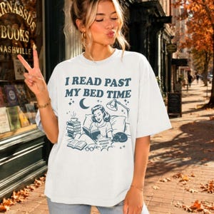 以下が含まれることがあります： I READ PAST MY BED TIMEというテキストが書かれたダークブルーのグラフィックプリントが特徴の白い半袖Tシャツ。イラストには、本の山、ランプ、三日月、猫と一緒にベッドで読書をする人物が描かれており、MIDNIGHT READERS CLUBというラベルが付いています。