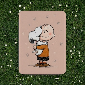 Op de afbeelding: Een roze tablethoes met een cartoonillustratie van Charlie Brown die Snoopy omhelst. Het ontwerp bevat kleine hartvormen verspreid over de achtergrond. De personages dragen hun klassieke outfits, Snoopy in wit en zwart en Charlie Brown in oranje en zwart.