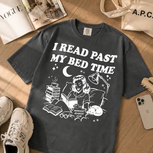 May include: A charcoal grey short-sleeve t-shirt featuring a white graphic print of a person reading in bed with a stack of books, a lamp, a cat, and the text "I READ PAST MY BED TIME" and "MIDNIGHT READERS CLUB" printed on the front chest area.