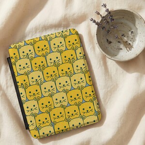 May include: A yellow cat-themed tablet case with a repeating pattern of smiling cat faces. The case has a black spine and is open, revealing a light blue interior. A small ceramic bowl with lavender sprigs sits nearby.