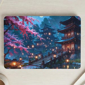 May include: A laptop skin featuring a serene Japanese garden scene. The design showcases traditional pagodas, a wooden bridge, and vibrant pink cherry blossoms. Lanterns illuminate the path, creating a tranquil and picturesque landscape.