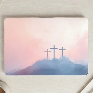 May include: Laptop skin featuring a watercolor design of three crosses on a hill. The background has a soft gradient of pink and peach hues, with a touch of blue at the base of the hill. The crosses are dark blue.