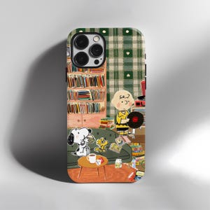 Op de afbeelding: Een smartphonehoesje met een Peanuts-illustratie van Charlie Brown die een vinylplaat vasthoudt, Snoopy die een sandwich eet en Woodstock op een groene bank. De achtergrond toont een boekenplank, een platenspeler en een groen ruitjespatroon.
