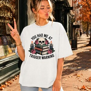 Puede incluir: Una camiseta blanca de manga corta con un estampado gráfico de una calavera sobre una pila de tres libros, flanqueada por dos velas encendidas y rosas rosadas, con el texto negro "YOU HAD ME AT TRIGGER WARNING" arqueado alrededor.