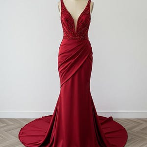 May include: A long, red, sleeveless gown with a plunging V-neckline and a beaded bodice. The gown has a fitted silhouette and a flowing train.