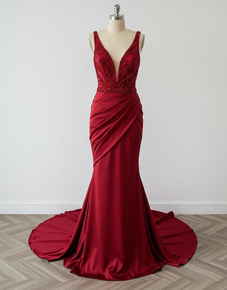 May include: A long, red, sleeveless gown with a plunging V-neckline and a beaded bodice. The gown has a fitted silhouette and a flowing train.
