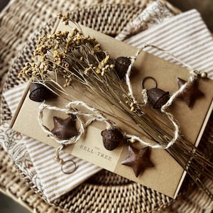 May include: A rectangular cardboard box labelled THE BELL TREE, topped with a bundle of dried yellow flowers and a rustic twine garland featuring dark brown metal acorn and star-shaped ornaments. The items are arranged on a flat surface.