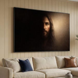 Puede incluir: Un retrato enmarcado de un hombre con pelo largo y barba, iluminado desde la derecha sobre un fondo oscuro. La obra de arte se exhibe sobre un sofá beige con cojines decorativos.